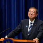 Shigeru Ishiba announcing his resignation as Prime Minister of Japan in front of a Japanese flag and microphones at a podium.
